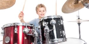 The image shows a smiling boy sitting behind a drum set, holding drumsticks raised in the air. The background is bright.